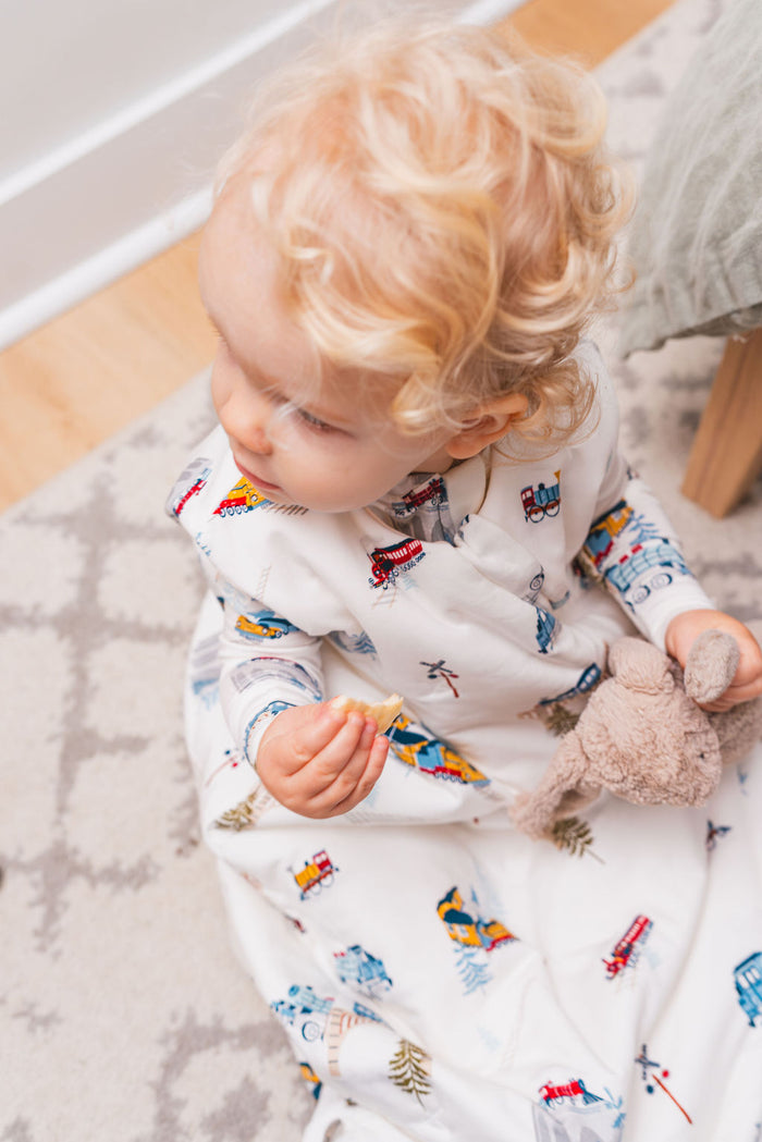 Child wearing a white onesie with a colorful train pattern, sitting on a wooden floor.