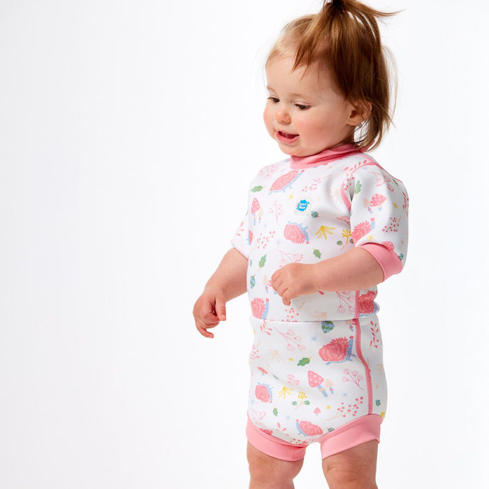 Lifestyle image of child wearing a baby wetsuit with built in swim diaper in white with pink trims and forest themed print, including hedgehogs, mushrooms and leaves. Side.