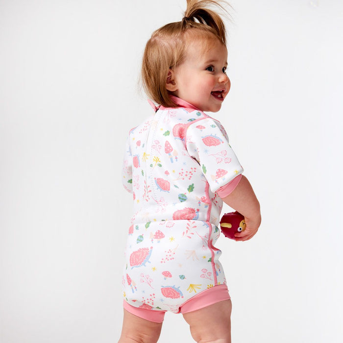 Lifestyle image of child wearing a baby wetsuit with built in swim diaper in white with pink trims and forest themed print, including hedgehogs, mushrooms and leaves. Back.