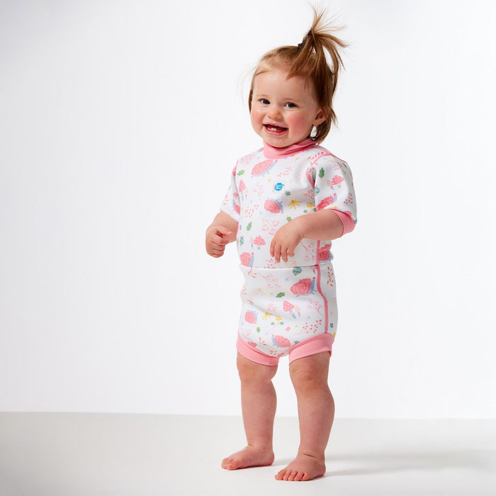 Lifestyle image of child wearing a baby wetsuit with built in swim diaper in white with pink trims and forest themed print, including hedgehogs, mushrooms and leaves. Front.