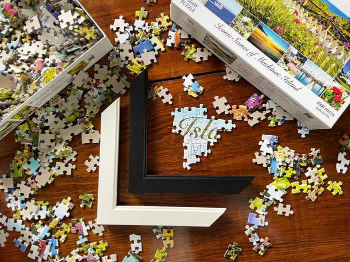 Jigsaw puzzle pieces on a wooden surface with a partially completed puzzle, Iconic Scenes of Mackinac Island puzzle box, and two frame samples.