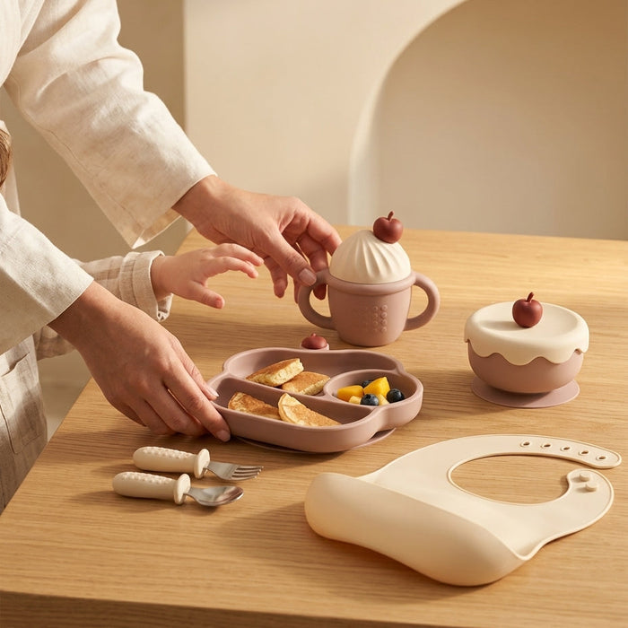 A styled tabletop scene showing a baby feeding set in warm neutral tones. A person in a cream linen outfit is placing a pastel pink divided plate with small food pieces into a small pink handled cup decorated like a dessert with a cherry on top.