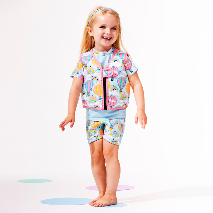 Lifestyle image of child wearing neoprene swim vest for toddlers with non-removable floats in baby blue, pink trims and hot air balloons themed print, including clouds and rainbows. She's also wearing matching jammers and rash top.