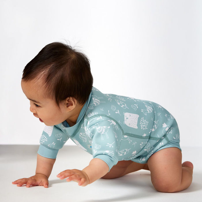 Lifestyle image of child wearing a baby wetsuit with built in swim diaper in greenish blue with animals themed print, including bears, birds and llamas. Side.