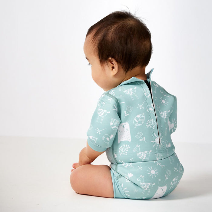 Lifestyle image of child wearing a baby wetsuit with built in swim diaper in greenish blue with animals themed print, including bears, birds and llamas. Back.