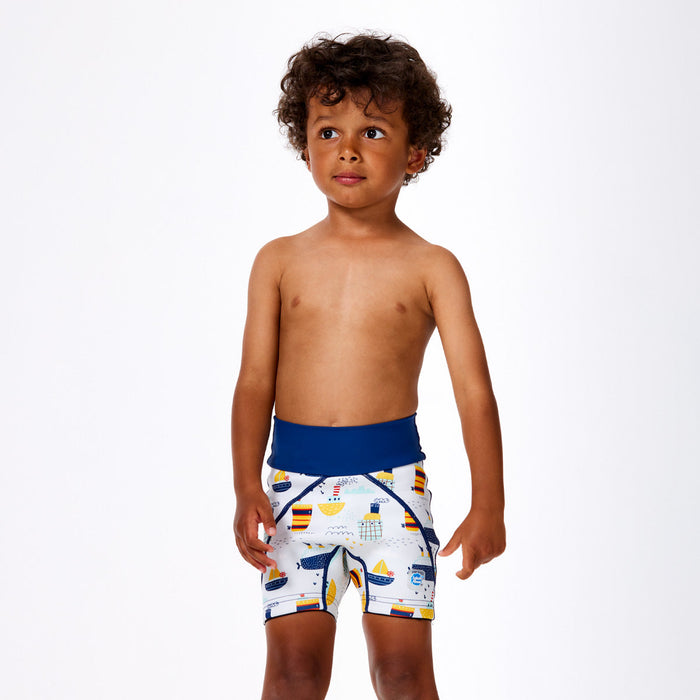 Little one wearing neoprene swim shorts in white with blue navy waist and boats themed print. Front.