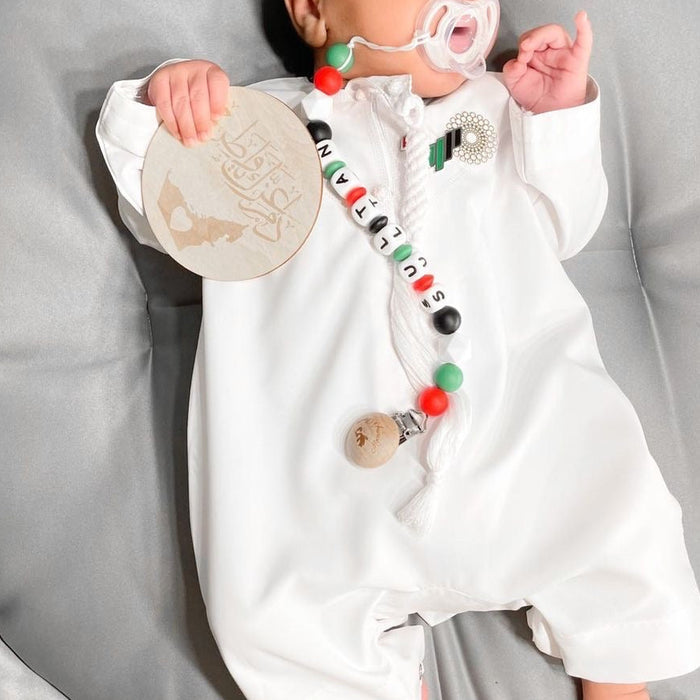 A baby wearing a white long-sleeve romper with colorful bead accents and a UAE NATIONAL DAY FLAG  pacifier clip, lying on a gray padded bassinet cushion.