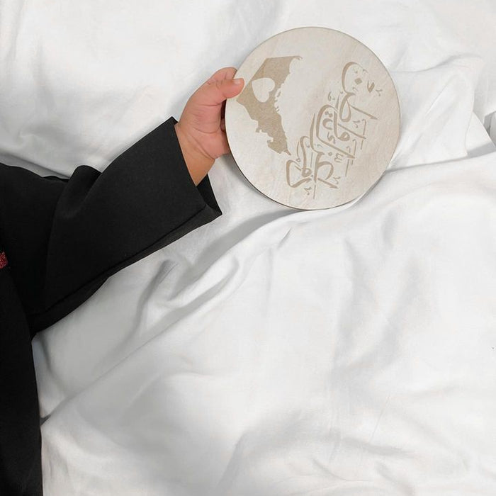 A hand holding a round ceramic plate with a beige map of EMIRATI and Arabic calligraphy, set against a background of white bedding.