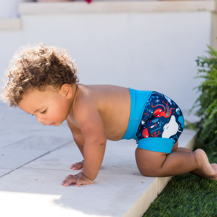 Child wearing reusable swim diaper in navy with cyan ribs and sea creatures themed print. Side.