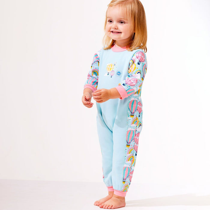 Lifestyle image of toddler wearing a fleece-lined baby wetsuit in baby blue with pink trims and hot air balloons themed print, including rainbows and clouds.