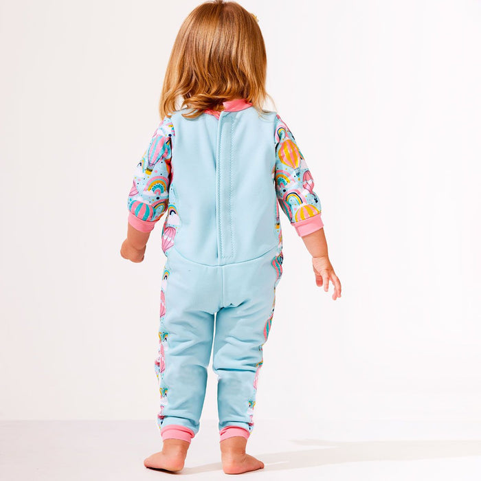 Lifestyle image of toddler wearing a fleece-lined baby wetsuit in baby blue with pink trims and hot air balloons themed print, including rainbows and clouds.